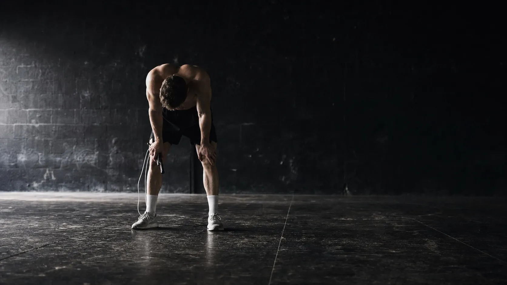 An athlete standing still between rounds in a dark training space, bent over with hands on knees and a rope hanging loose from one hand.