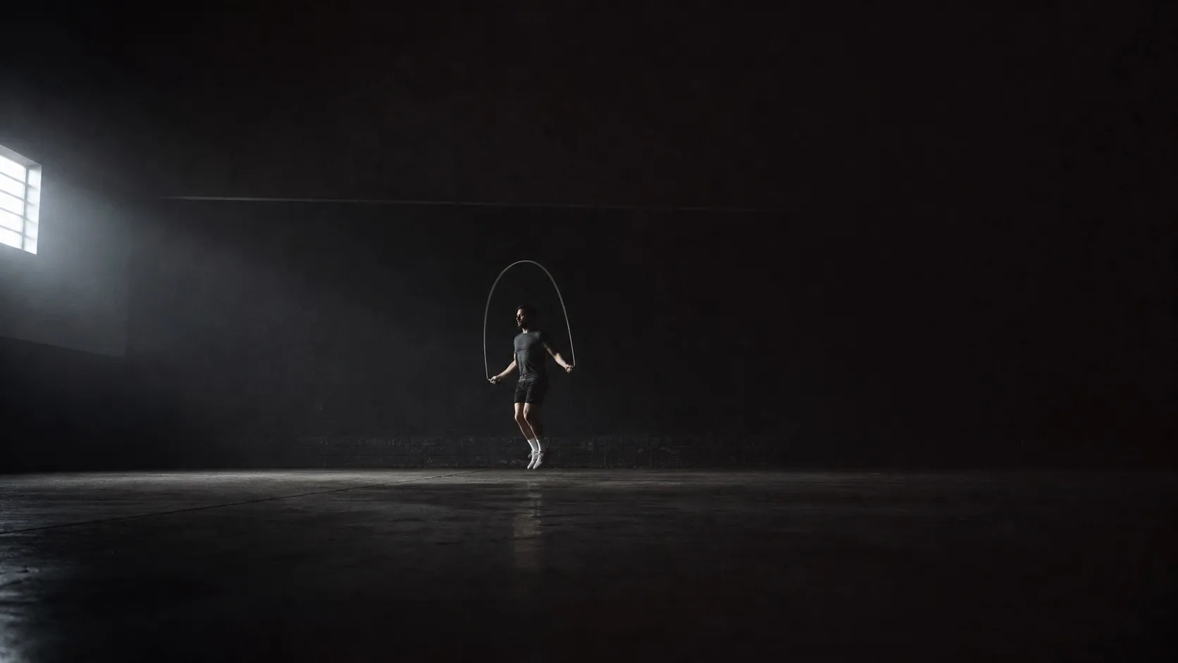 A lone athlete skipping in a dark, almost empty training space, the rope caught as a blurred arc under one hard side light.
